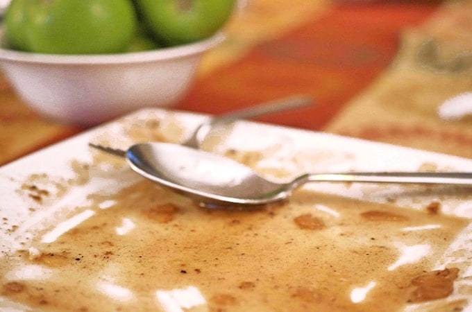 Plate of remaining sauce holding 2 serving utensils, bowl of green apples in background