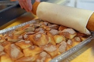 Aluminum pan holding prepped apples being covered by dough on rolling pin