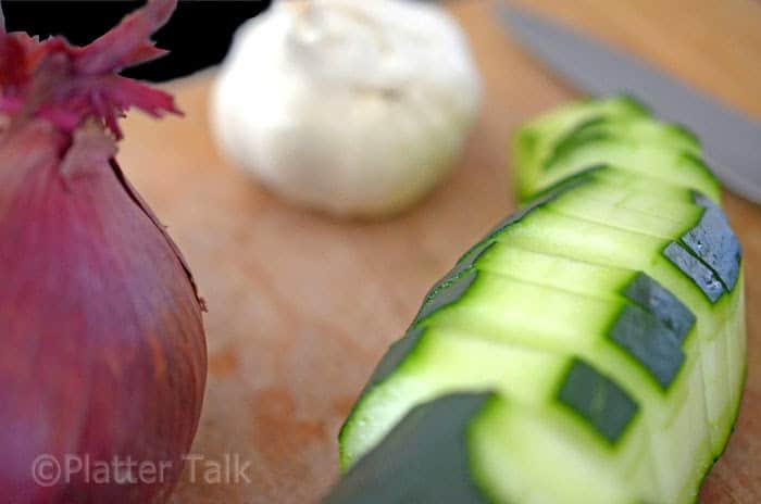 A close up of a garlic, red onion and sliced cuke on cutting board with knife