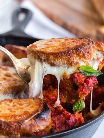 Scooping a portion of meatballs and bread out of skillet with melted cheese.