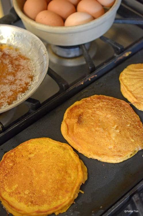 Pumpkin pancakes on a kitchen griddle with eggs in the background.