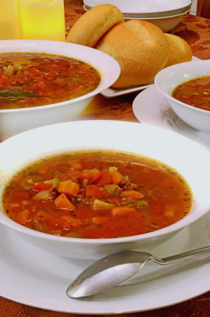 Bowl of hamburger soup with dinner rolls in the bacground.