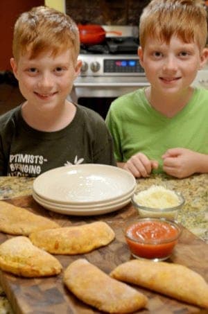 2 boys behind wood board, plates, calzones, tomato sauce and grated cheese