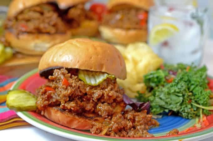 A plate of Sloppy Joe, chips and salad in front of 3 Joes on a board