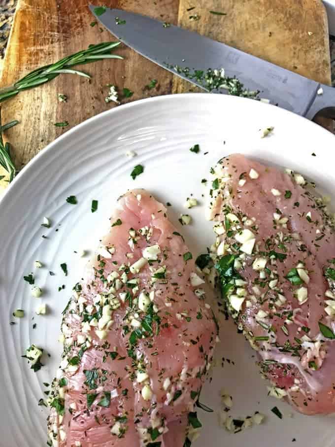 Two chicken breasts seasoned with fresh herbs, on a cutting board.