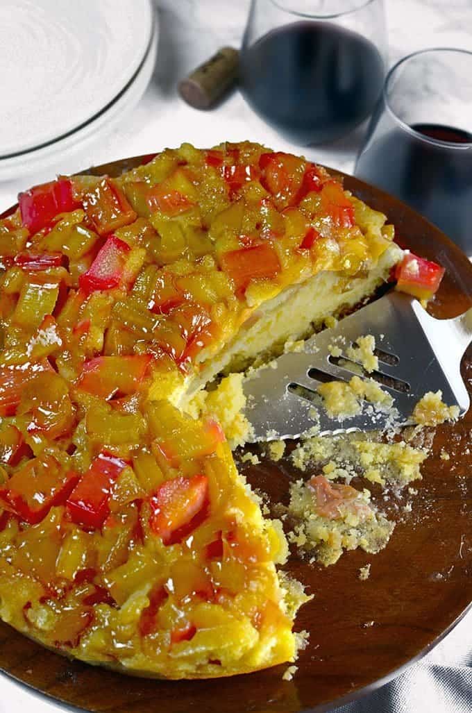 Rhubarb cake and spatula on a cake pedistal with wine glasses in background.