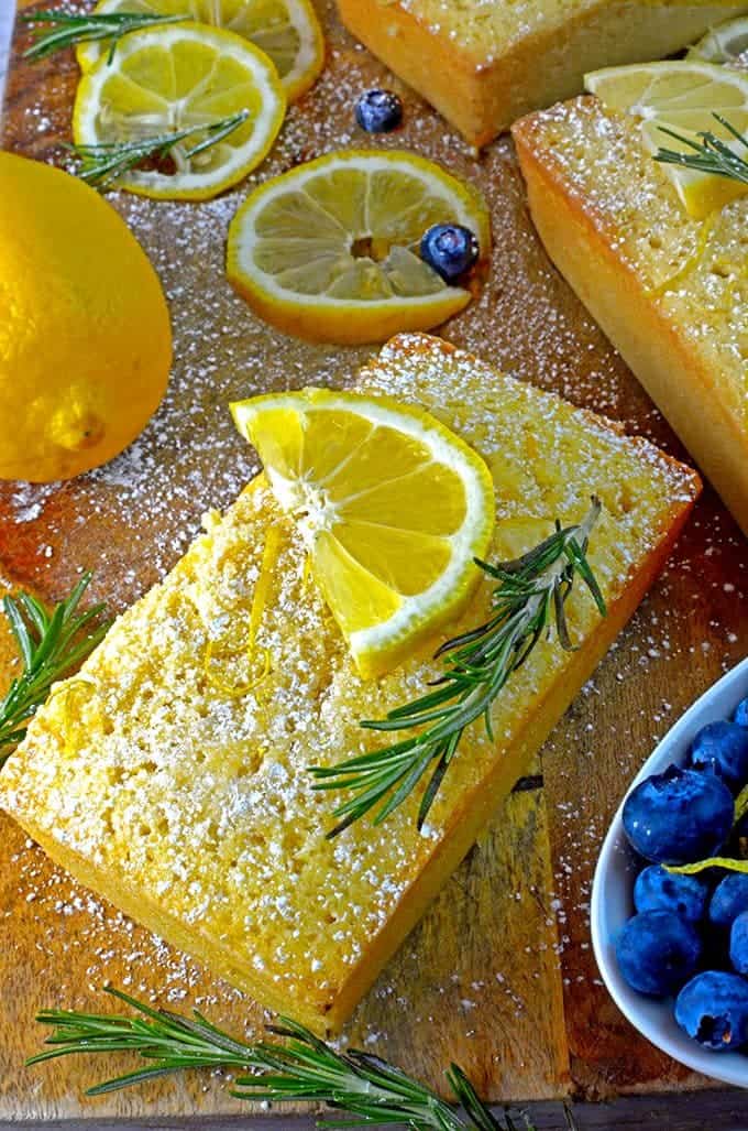 Lemon loaf cake with slices of oragne and sprigs of rosemary on a cutting board.