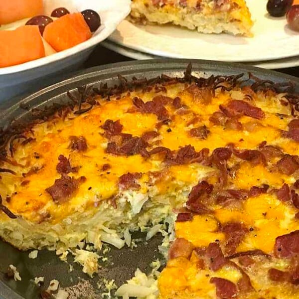 Serving dish of hash brown quiche with bacon, plated slice with fruit in back