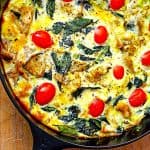 Close up of iron skillet containing batter, tomato, greens on top of wood