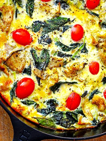 Close up of iron skillet containing batter, tomato, greens on top of wood