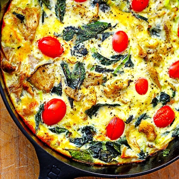 Close up of iron skillet containing batter, tomato, greens on top of wood