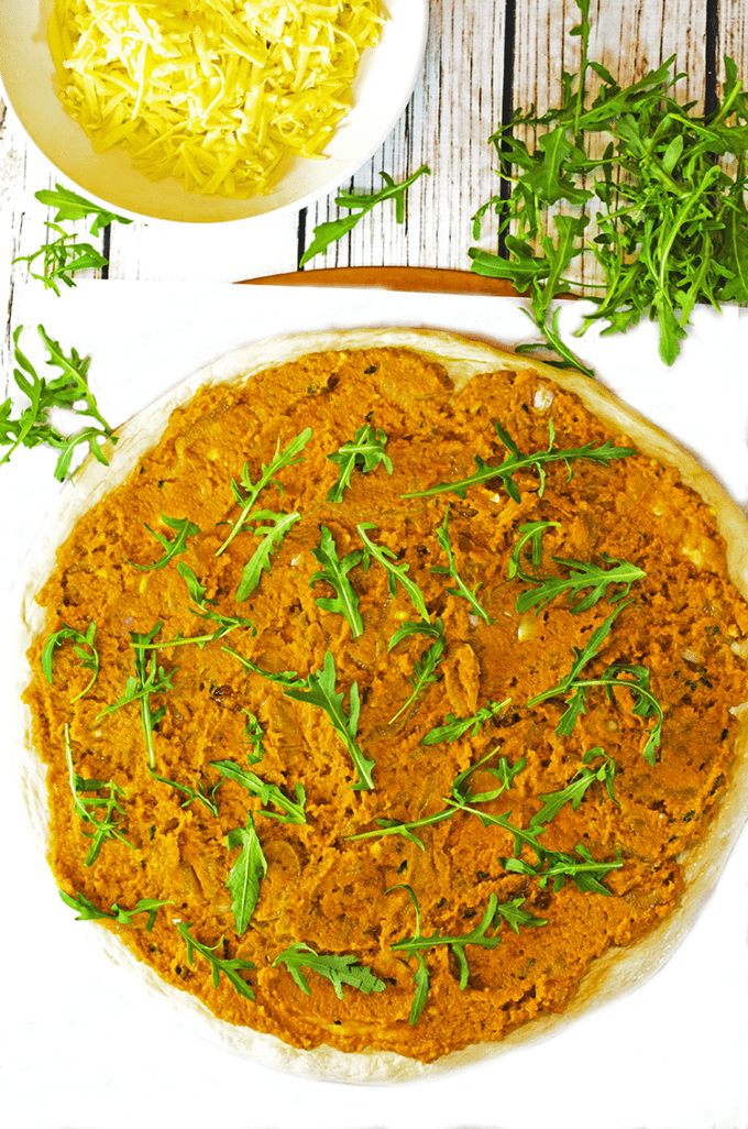 Cooked pumpkin mixture and fresh arugula atop raw pizza resting on cloth 