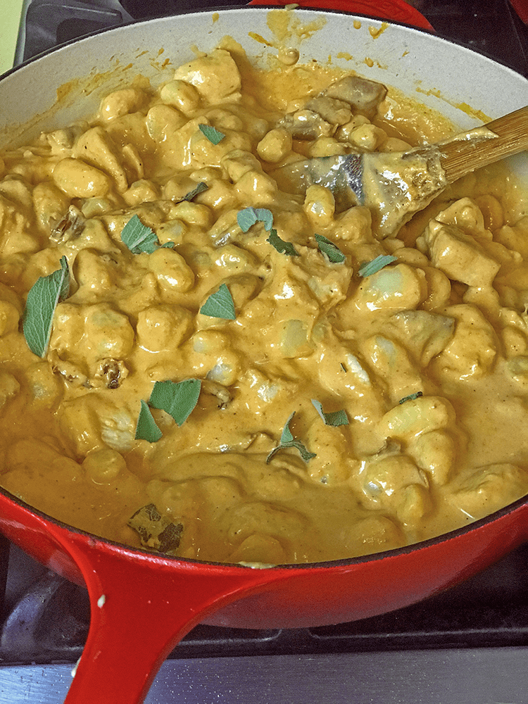 Pot of cooking, sauced pumpkin gnocchi being stirred with fresh herbs using wood spoon