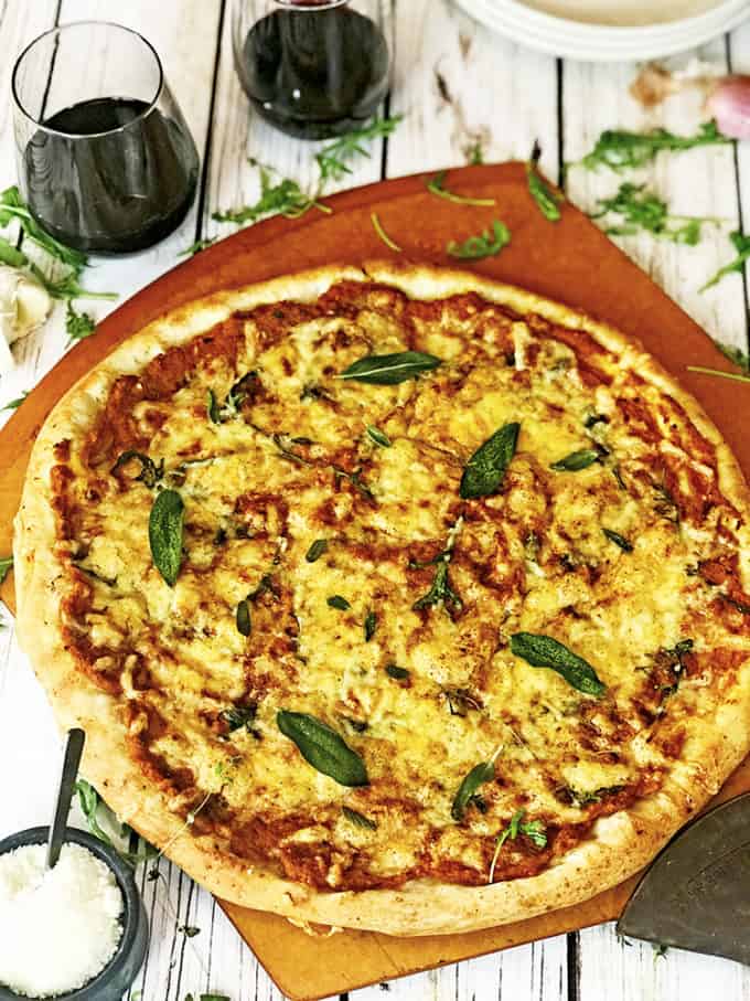 Freshly baked uncut pumpkin pizza with sage atop a wooden board and table 