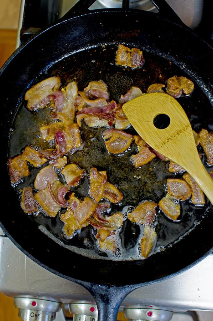Cast iron skillet with bacon pieces being fried and a wooden sppon.