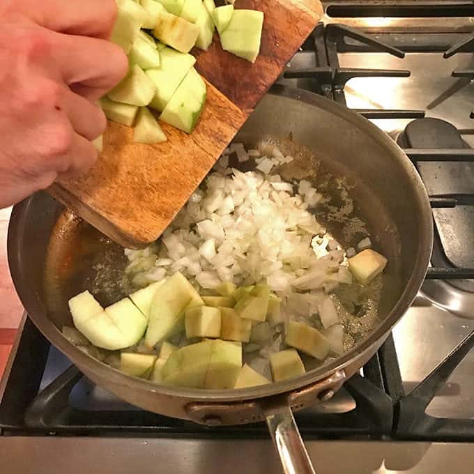 Someone adding apples from cutting board to pan of onions on stove