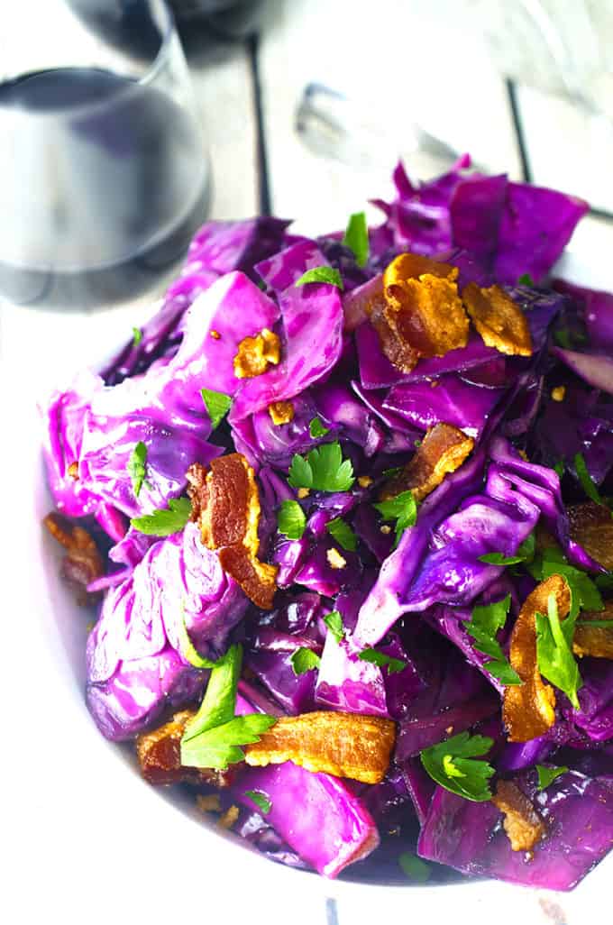 Bowl of purple fried cabbage with bacon and parsely garnsih and a glass of wine.