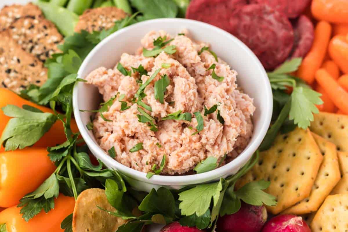 A bowl of ham salad surrounded by crackers and vegetables.