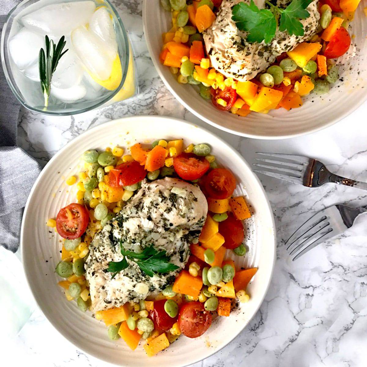 Two bowls of succotash topped with chicken breast and a glass of ice water.