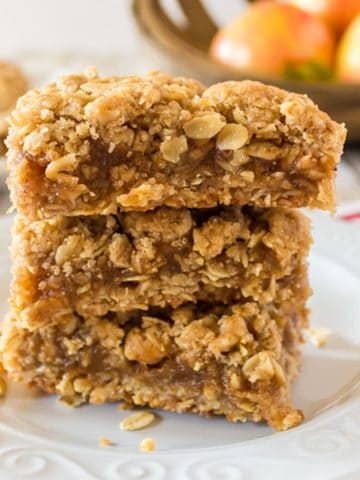 stack of three oatmeal dessert bars with appeles in the background,.