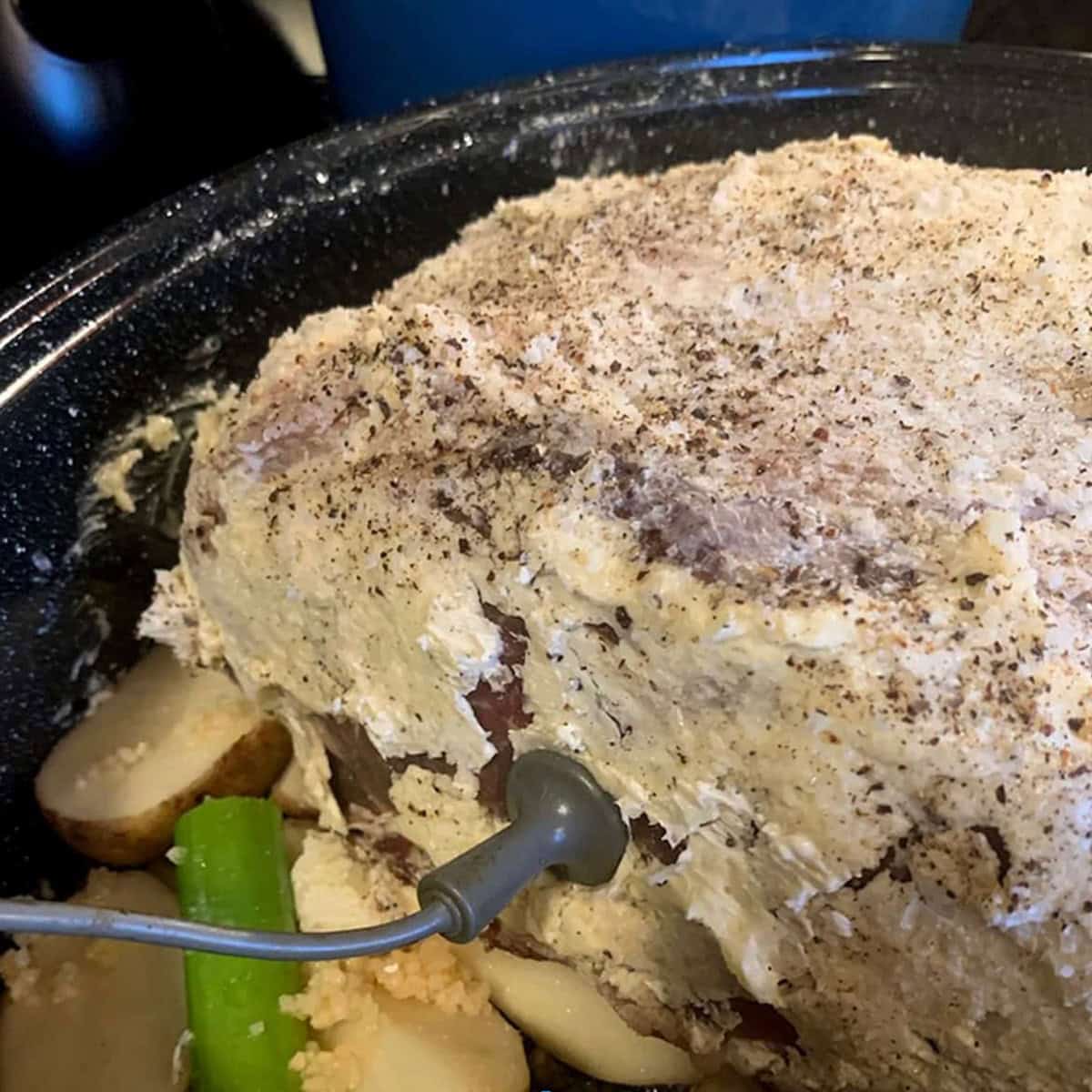 A hunk of prime rib covered in butter sitting in a roaster with a temp probe in it.