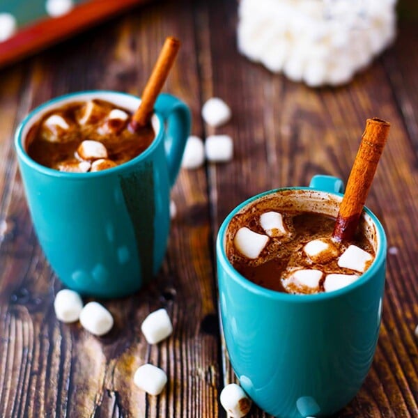 Two blue mugs of vegan hot chocolate with marshmallows and cinnamon sticks.
