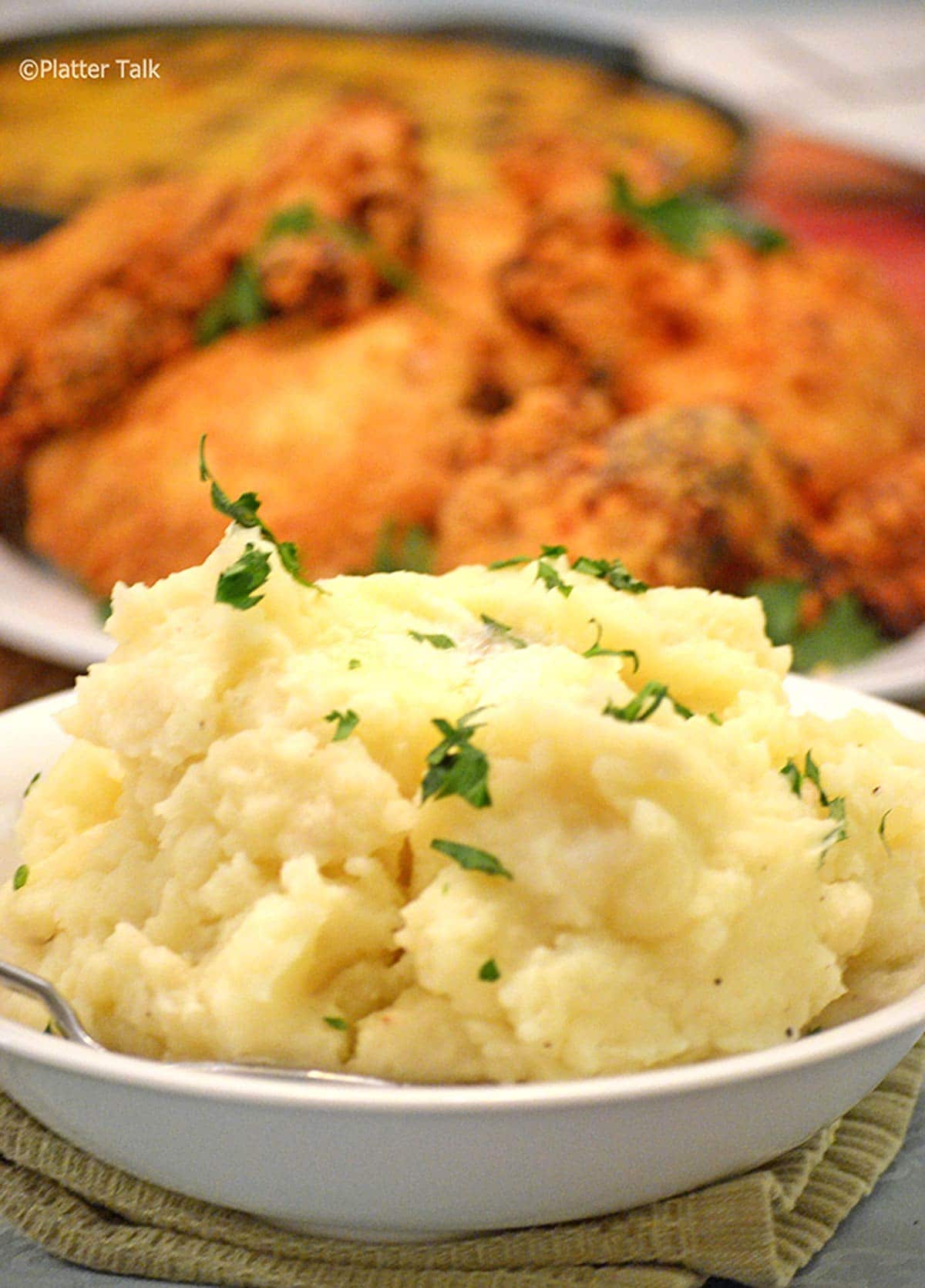 A bowl of mashed potatoes on a table with fried chicken in background.