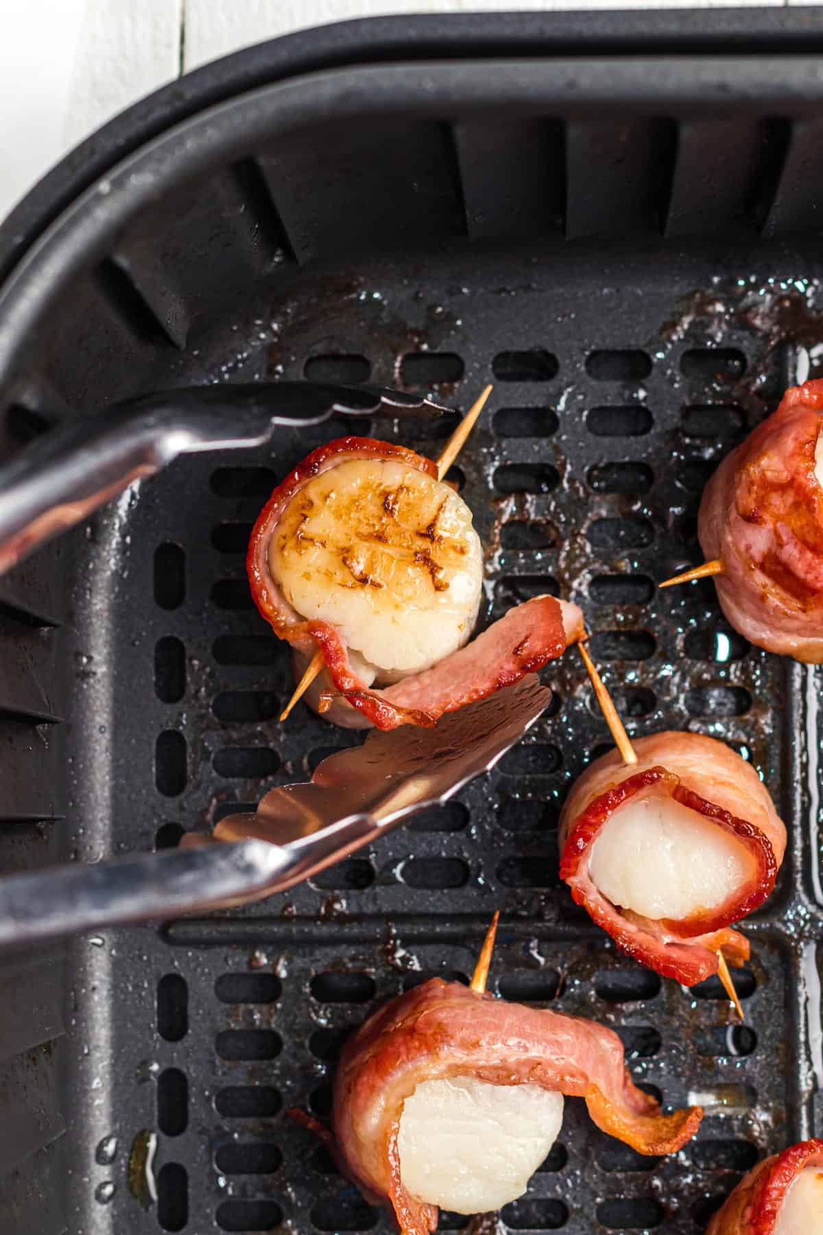 Removing bacon-wrapped scallops from an air fryer using tongs.