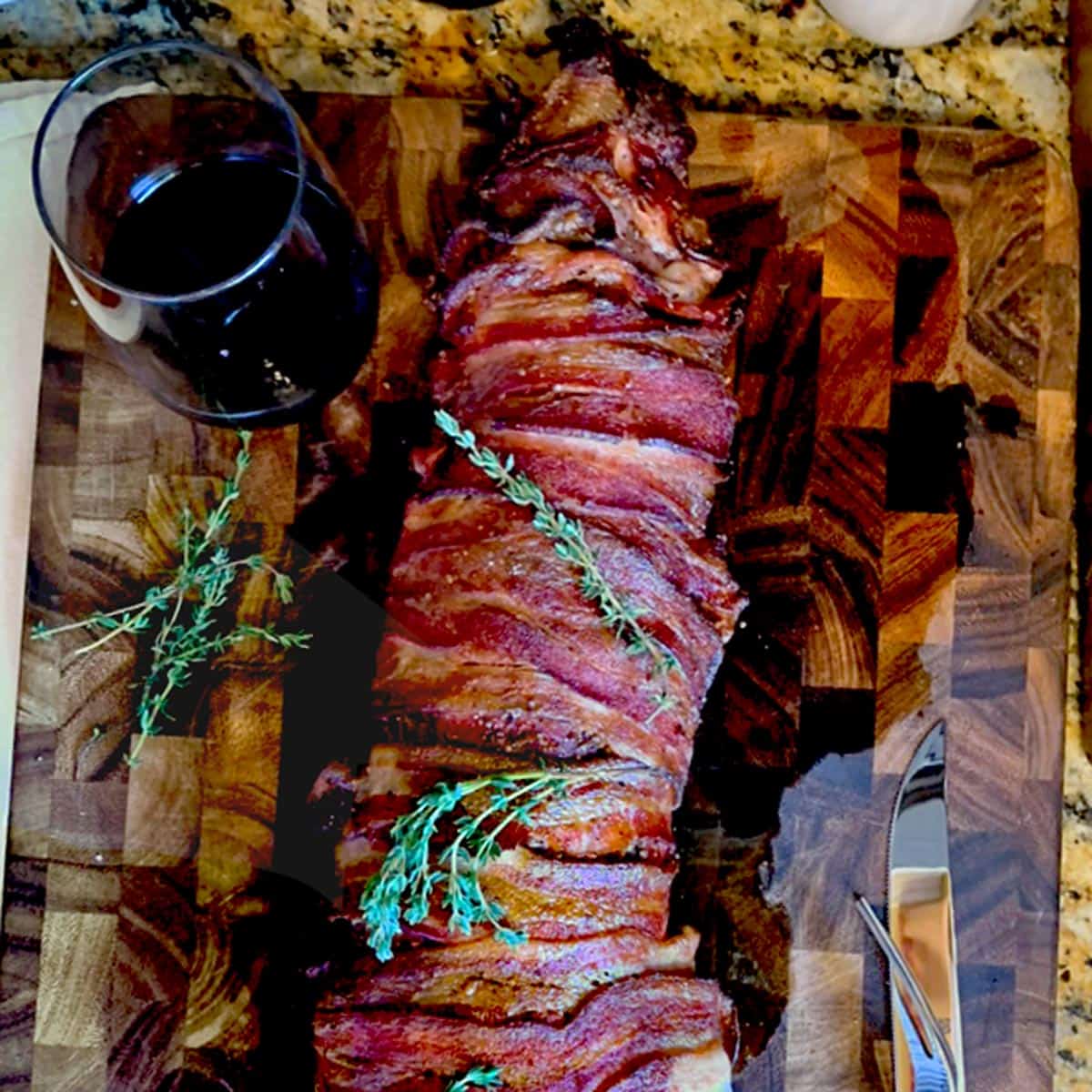 A whole beef tenderloin roast wrapped in bacon and sitting on a cutting board.