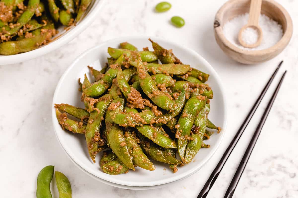 A bowl of garlic edamame pods with a pair of chop sticks.