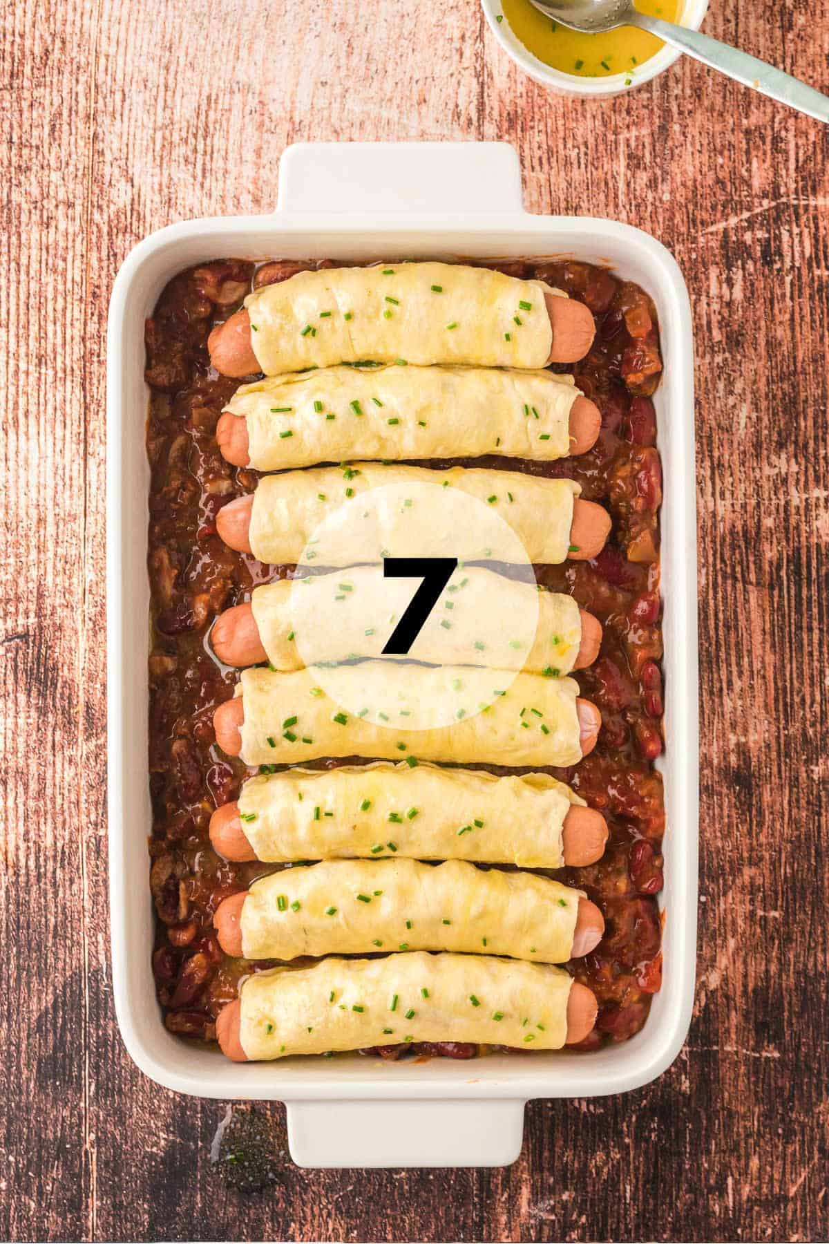 A bunch of hot dogs wrapped in dough in a pan of chili.