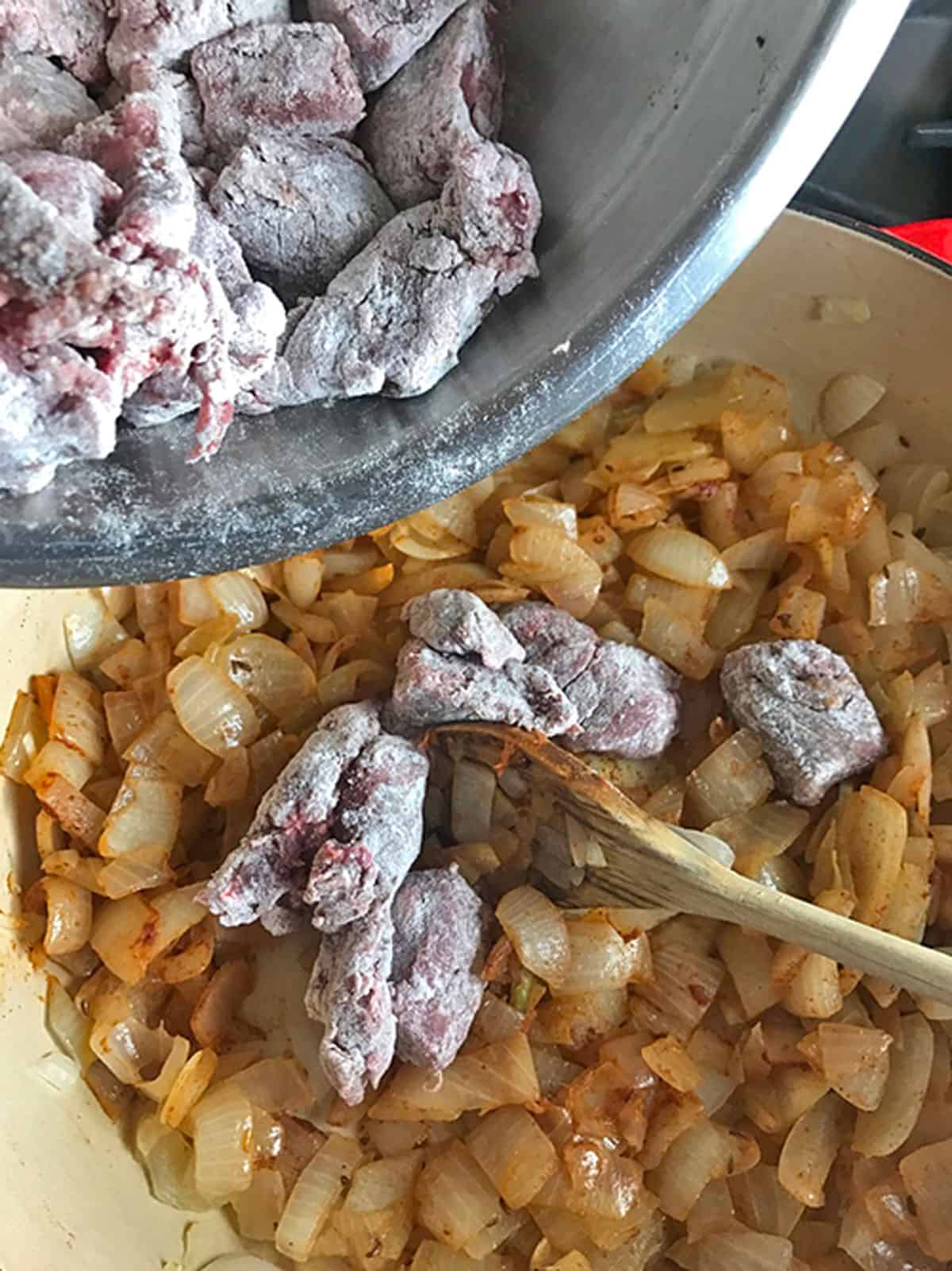 Some flour-coated chunks of beef being mixed into a pot of cooked onions.