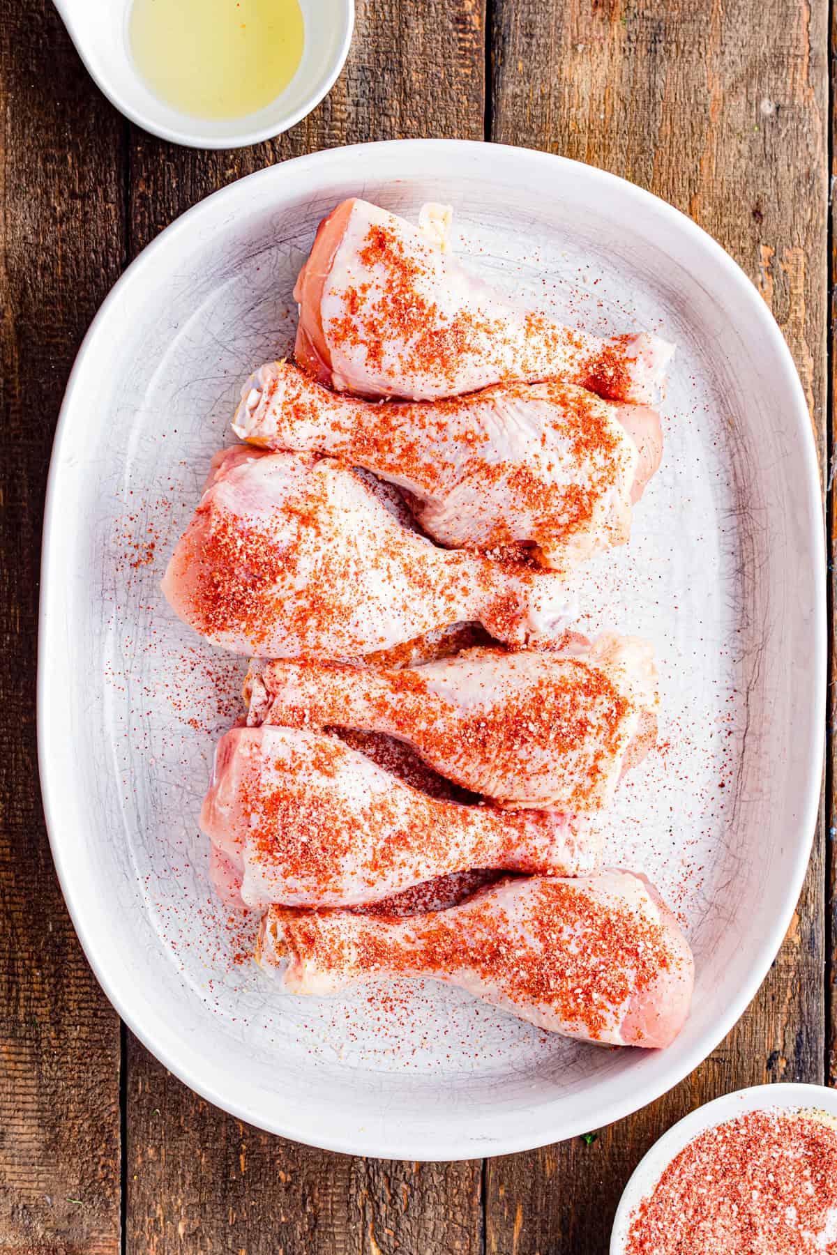 A plate of uncooked chicken drumsticks with seasoning sprinkled on them.