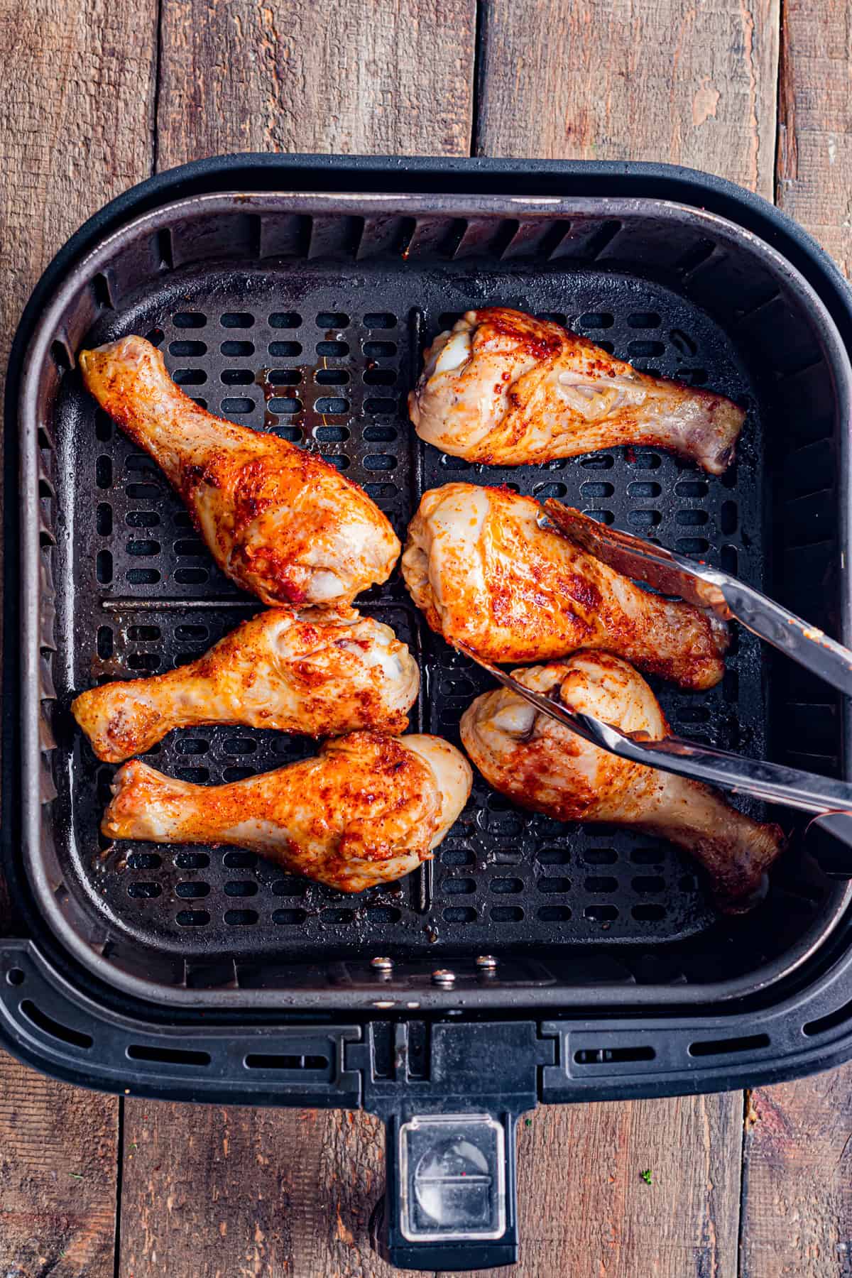 A kitchen tongs, turning some air fryer chicken drumsticks, midway through cooking.